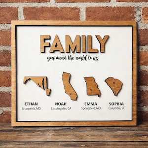 May include: Wooden family sign with the word "FAMILY" in raised letters above the phrase "you mean the world to us." Four state outlines are below with names and locations: Ethan, Noah, Emma, and Sophia. The sign is framed.
