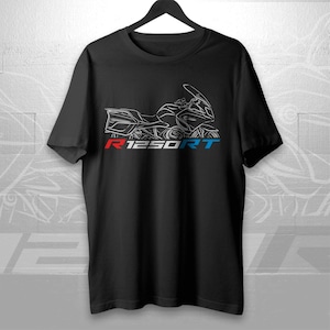 Puede incluir: Camiseta negra con un contorno blanco de una motocicleta y el texto "R1250RT" en rojo, blanco y azul. La camiseta está colgada en una percha negra sobre un fondo blanco.
