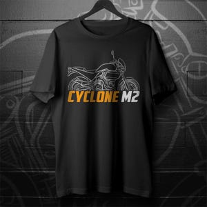 Può includere: Una t-shirt nera a maniche corte con un'illustrazione a linee bianche di una motocicletta centrata sul petto. Sotto la grafica della moto, il testo CYCLONE M2 è stampato in grassetto, in lettere maiuscole arancioni e bianche sul davanti.