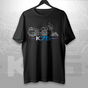 May include: Black t-shirt featuring a white line drawing of a motorcycle with saddlebags and a windshield. The text "K75" is in white, with red and blue lines above and below the text. The shirt is hanging on a black hanger.