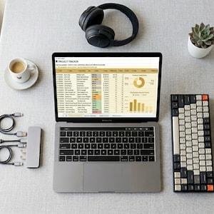 May include: A laptop displaying a project tracker spreadsheet, with a black keyboard, headphones, and a small succulent plant. Also present are a coffee cup, a notebook, a pen, and a USB hub with cables. The setup suggests a workspace or home office.