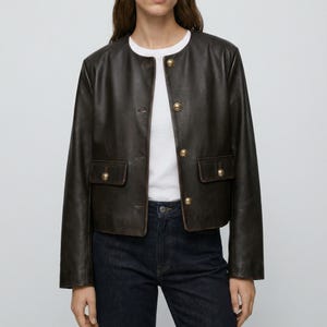 以下が含まれることがあります： 丸襟と金色のボタンが付いた、濃い茶色のレザージャケット。 ジャケットには、お揃いの金色のボタンが付いた2つのフロントポケットがあります。 モデルは白いTシャツと濃い青色のジーンズを着用しています。