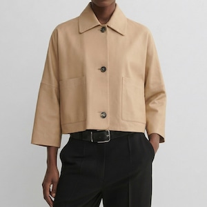 May include: A tan cropped jacket with a collar, three dark buttons, and two front pockets. The jacket has three-quarter sleeves and is worn with black pants and a black belt.