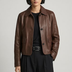 May include: A brown leather jacket with a collared neckline, two front pockets, and button closures. The jacket is worn over a black top and paired with black pants. A black belt with a silver buckle is visible.