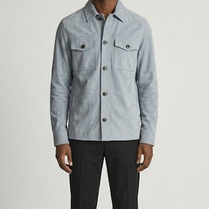 May include: A light blue suede shirt jacket with two chest pockets and button closures. The jacket has a classic collar and long sleeves. The model is wearing black pants.