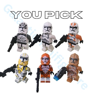 May include: A collection of six Lego minifigures of clone troopers, each with unique helmet designs and armor markings. The figures are in various colors, including white, orange, yellow, and brown, and are holding black blasters. The text "YOU PICK" is at the top.