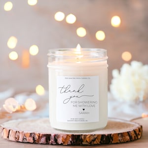 Puede incluir: Una vela blanca de soja en un tarro de vidrio transparente con una etiqueta que dice "THE DANCING WICK CANDLE CO. thank you FOR SHOWERING ME WITH LOVE SARAH PURE SOY CANDLE HANDPOURED IN BIXBY, OK"