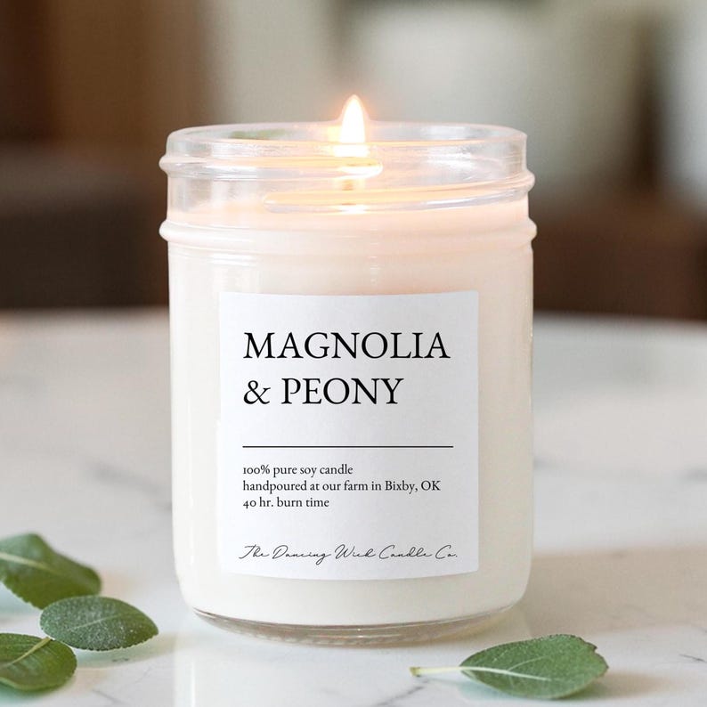 May include: A lit soy candle in a clear glass jar with a white label reading Magnolia & Peony, 100% pure soy candle, handpoured at our farm in Bixby, OK, 40 hr. burn time, and The Dancing Wick Candle Co. The candle has a single burning wick.
