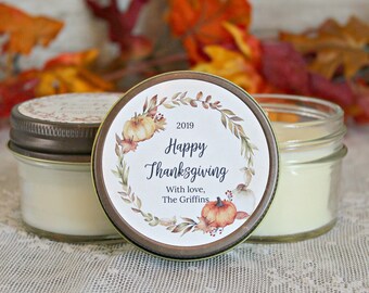 May include: A white candle in a glass jar with a brown lid. The lid has a round sticker with a watercolor design of fall leaves, pumpkins, and the text "2019 Happy Thanksgiving With love, The Griffins".