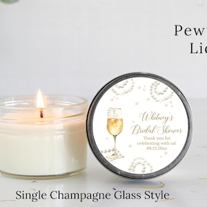 May include: A white candle in a clear glass jar with a pewter lid. The lid has a white label with gold text that reads "Whitney's Bridal Shower" and "Thank you for celebrating with us! 08.13.20xx". The label also features a watercolor illustration of a champagne flute with pearls.