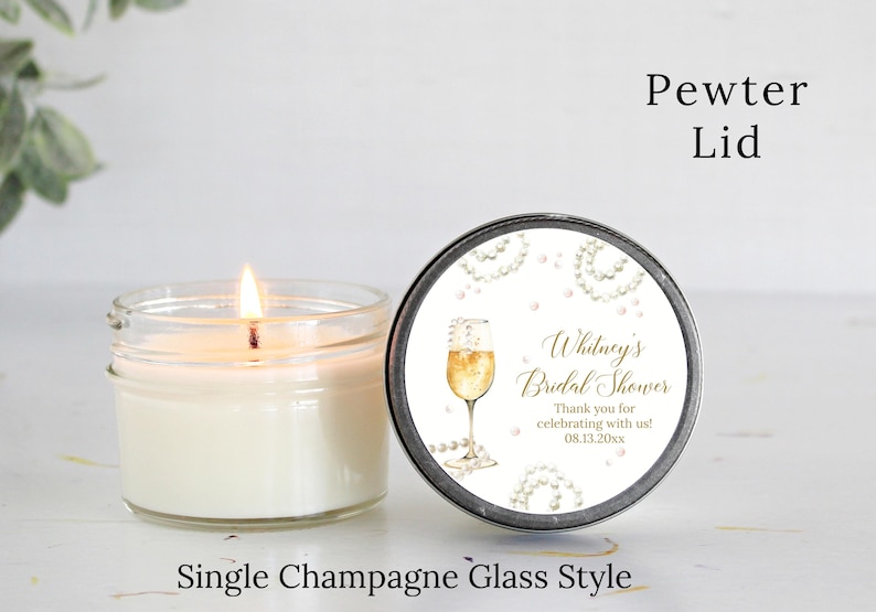 May include: A white candle in a clear glass jar with a pewter lid. The lid has a white label with gold text that reads "Whitney's Bridal Shower" and "Thank you for celebrating with us! 08.13.20xx". The label also features a watercolor illustration of a champagne flute with pearls.