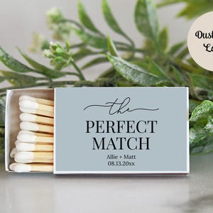 Peut inclure: Une bo&icirc;te d'allumettes rectangulaire avec une &eacute;tiquette bleu poussi&eacute;reux portant le texte "the PERFECT MATCH" en noir, ainsi que les noms "Allie + Matt" et la date "08.13.20xx". La bo&icirc;te contient des allumettes en bois &agrave; bouts blancs.