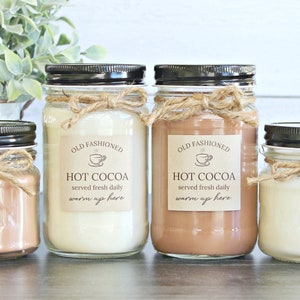 May include: Four mason jar candles with brown lids and twine bows. The labels read "Old Fashioned Hot Cocoa served fresh daily warm up here".