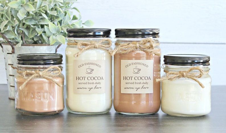 May include: Four mason jar candles with brown lids and twine bows. The labels read "Old Fashioned Hot Cocoa served fresh daily warm up here".