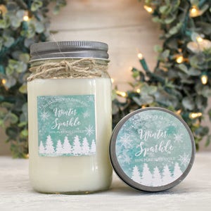 May include: A glass jar candle with a silver lid and a white label that says "Winter Sparkle" and "100% Pure Soy Candle". The label has a blue and green watercolor background with snowflakes and pine trees.