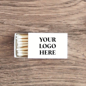 Pu&ograve; includere: Una scatola di fiammiferi bianca con un'etichetta bianca che dice "YOUR LOGO HERE" e una fila di fiammiferi con teste bianche e stecchi marroni.