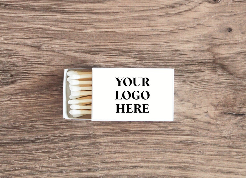 Pu&ograve; includere: Una scatola di fiammiferi bianca con un'etichetta bianca che dice "YOUR LOGO HERE" e una fila di fiammiferi con teste bianche e stecchi marroni.