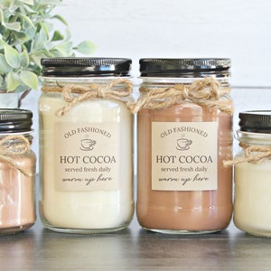 May include: Four glass mason jars with black lids and twine bows. The jars contain a brown, white, and beige substance. Each jar has a label that reads "Old Fashioned Hot Cocoa served fresh daily warm up here".