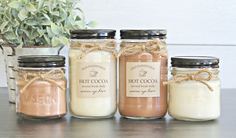 May include: Four glass mason jars with black lids and twine bows. The jars contain a brown, white, and beige substance. Each jar has a label that reads "Old Fashioned Hot Cocoa served fresh daily warm up here".
