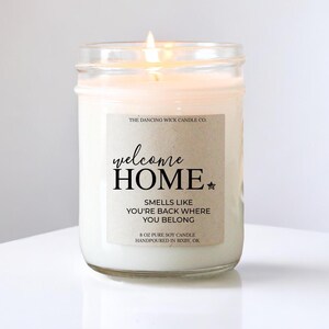May include: A white soy candle in a glass jar with a brown label that reads "welcome HOME", "SMELLS LIKE YOU'RE BACK WHERE YOU BELONG", "8 OZ PURE SOY CANDLE HANDPOURED IN BIXBY, OK" and "THE DANCING WICK CANDLE CO."