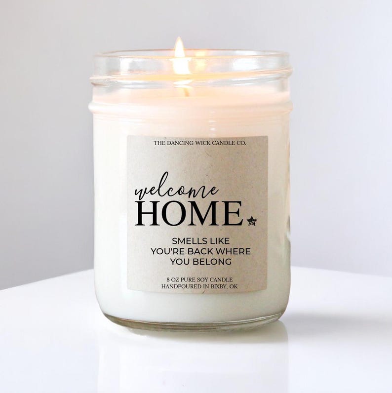 May include: A white soy candle in a glass jar with a brown label that reads "welcome HOME", "SMELLS LIKE YOU'RE BACK WHERE YOU BELONG", "8 OZ PURE SOY CANDLE HANDPOURED IN BIXBY, OK" and "THE DANCING WICK CANDLE CO."