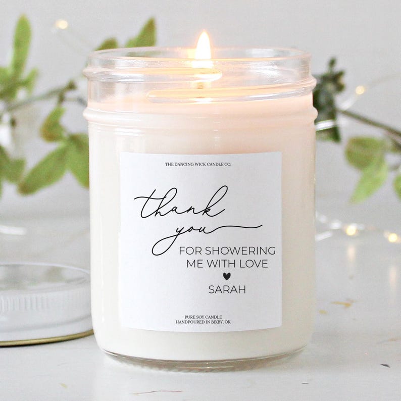 Puede incluir: Una vela de soja blanca en un tarro de vidrio transparente con una etiqueta blanca que dice "The Dancing Wick Candle Co." y "Gracias por llenarme de amor Sarah".