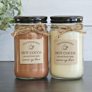 May include: Two glass jars with black lids and twine tied around the top. The jars are filled with a brown and white substance. The labels on the jars read "Old Fashioned Hot Cocoa served fresh daily warm up here".