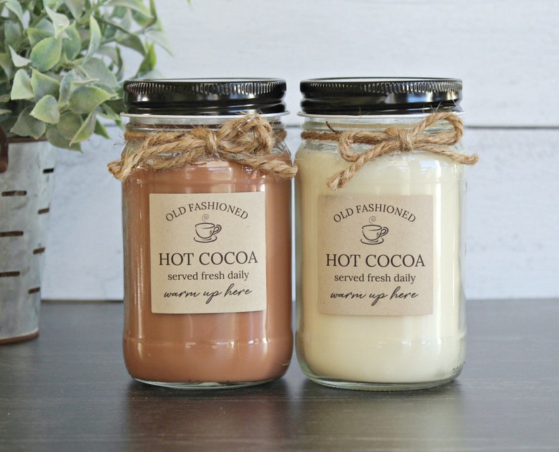 May include: Two glass jars with black lids and twine tied around the top. The jars are filled with a brown and white substance. The labels on the jars read "Old Fashioned Hot Cocoa served fresh daily warm up here".