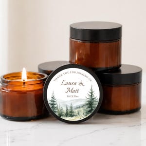 May include: Four amber glass jars with black lids, one lit candle, and a circular label featuring a watercolor forest landscape, the text "THANK YOU FOR JOINING US!", "Laura & Matt", and the date "10.13.20xx" centered on the front jar.