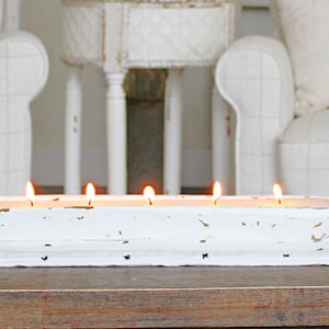 May include: A white wooden trough filled with a candle with five wicks, burning on a white table runner with black polka dots. The candle is in a white room with two white chairs and a white table in the background.