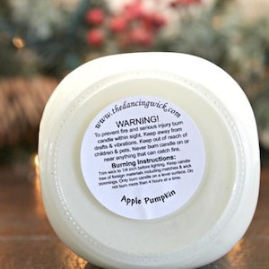 May include: A white candle in a glass jar with a circular label on the bottom. The label features the text www.thedancingwick.com, a warning about fire safety, burning instructions, and the scent name Apple Pumpkin in black text.