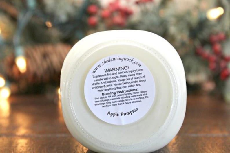 May include: A white candle in a glass jar with a circular label on the bottom. The label features the text www.thedancingwick.com, a warning about fire safety, burning instructions, and the scent name Apple Pumpkin in black text.