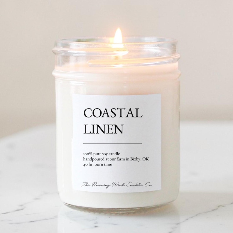 May include: A clear glass jar candle with a lit wick and off-white wax. The white label features the text COASTAL LINEN, 100% pure soy candle, handpoured at our farm in Bixby, OK, 40 hr. burn time, and The Dancing Wick Candle Co.