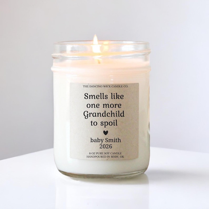 Puede incluir: Una vela blanca encendida en un tarro de cristal. La etiqueta dice "Smells like one more Grandchild to spoil" con el nombre "baby Smith" y el a&ntilde;o "2026". La vela es de 227g y vertida a mano.
