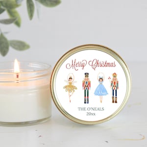 May include: A lit candle next to a round, gold-rimmed container with a festive design. The container features the words "Merry Christmas" and illustrations of a ballerina and nutcrackers. The text "The O'Neals" and a year are also included.