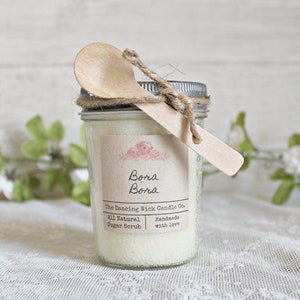 May include: A glass jar filled with white sugar scrub, tied with twine and a wooden spoon. The label reads "Bora Bora The Dancing Wick Candle Co. All Natural Sugar Scrub Handmade with Love."