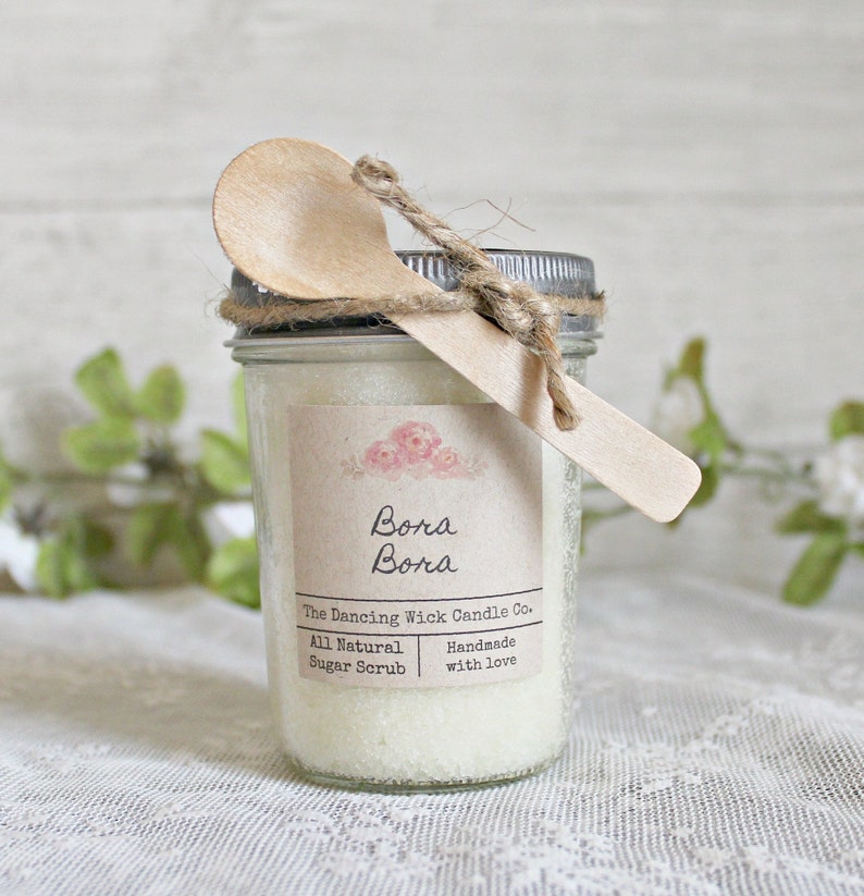 May include: A glass jar filled with white sugar scrub, tied with twine and a wooden spoon. The label reads "Bora Bora The Dancing Wick Candle Co. All Natural Sugar Scrub Handmade with Love."