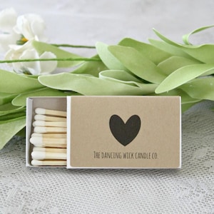 K&ouml;nnte beinhalten: Eine Schachtel Streichh&ouml;lzer mit einem braunen Papieraufkleber. Der Aufkleber hat ein schwarzes Herz und den Text "The Dancing Wick Candle Co."