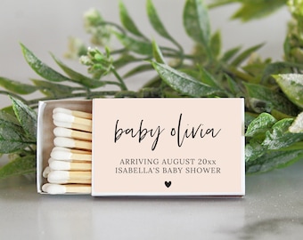 Op de afbeelding: Een roze luciferdoosje met een witte rand en een zwart hart. Het luciferdoosje heeft de tekst "baby olivia" in zwarte cursieve letters. De tekst "arriving august 20xx" en "isabella's baby shower" is ook in zwart gedrukt.