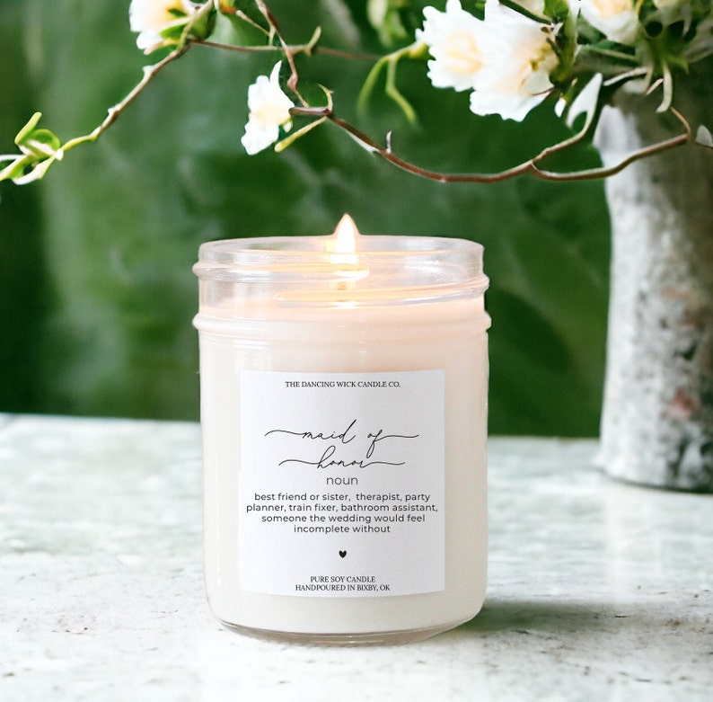 May include: A white candle in a clear glass jar with a label that reads "The Dancing Wick Candle Co. maid of honor noun best friend or sister, therapist, party planner, train fixer, bathroom assistant, someone the wedding would feel incomplete without Pure Soy Candle Handpoured in Bixby, OK"