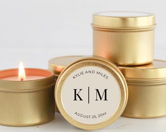 May include: Gold-colored candle tins, some stacked, with a lit candle in one. The labels read "Kylie and Miles" with the initials "K | M" and the date "August 25, 20xx". The candles are likely for a wedding or special event.