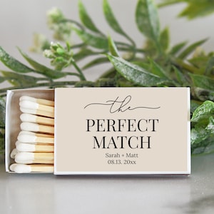 Peut inclure: Une bo&icirc;te d'allumettes rectangulaire avec une couverture beige clair portant le texte noir "the PERFECT MATCH" suivi de "Sarah + Matt" et "08.13. 20xx". La bo&icirc;te est entrouverte, r&eacute;v&eacute;lant une rang&eacute;e d'allumettes en bois &agrave; bouts blancs.