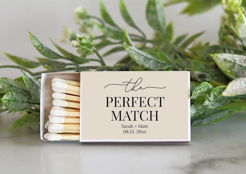 Peut inclure: Une bo&icirc;te d'allumettes rectangulaire avec une couverture beige clair portant le texte noir "the PERFECT MATCH" suivi de "Sarah + Matt" et "08.13. 20xx". La bo&icirc;te est entrouverte, r&eacute;v&eacute;lant une rang&eacute;e d'allumettes en bois &agrave; bouts blancs.