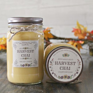 K&ouml;nnte beinhalten: Zwei Glasbeh&auml;lter mit braunen Deckeln und Etiketten. Die Etiketten zeigen "The Dancing Wick Candle Co." und "Harvest Chai 100% Soy Candle". Die Beh&auml;lter sind mit einem hellbraunen Wachs gef&uuml;llt.
