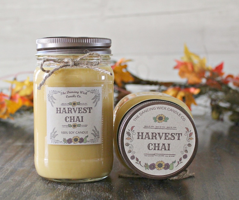 K&ouml;nnte beinhalten: Zwei Glasbeh&auml;lter mit braunen Deckeln und Etiketten. Die Etiketten zeigen "The Dancing Wick Candle Co." und "Harvest Chai 100% Soy Candle". Die Beh&auml;lter sind mit einem hellbraunen Wachs gef&uuml;llt.