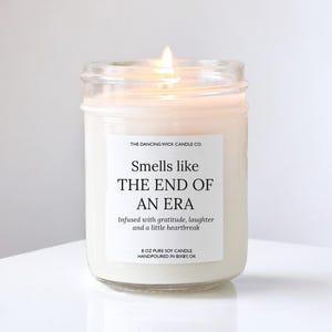 May include: A clear glass jar candle with a white label featuring the text: THE DANCING WICK CANDLE CO. Smells like THE END OF AN ERA Infused with gratitude, laughter and a little heartbreak 8 OZ PURE SOY CANDLE HANDPOURED IN BIXBY, OK. The wax is off-white.