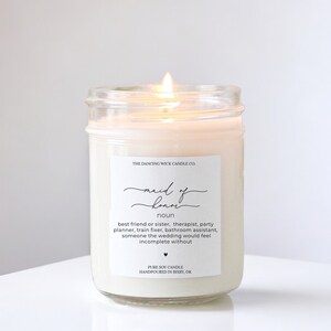 May include: A white candle in a clear glass jar with a label that reads "Maid of Honor" and defines the term as "best friend or sister, therapist, party planner, train fixer, bathroom assistant, someone the wedding would feel incomplete without." The label also states "Pure Soy Candle Handpoured in Bixby, OK."