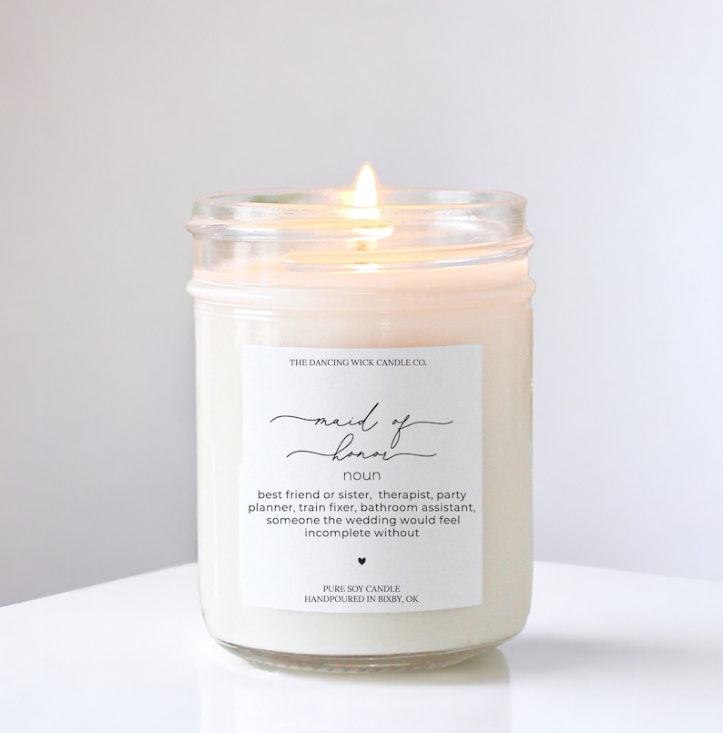 May include: A white candle in a clear glass jar with a label that reads "Maid of Honor" and defines the term as "best friend or sister, therapist, party planner, train fixer, bathroom assistant, someone the wedding would feel incomplete without." The label also states "Pure Soy Candle Handpoured in Bixby, OK."