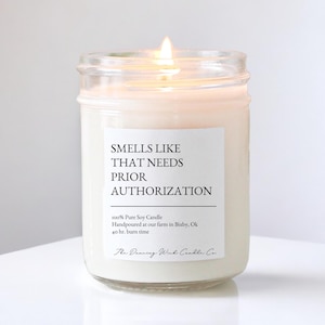May include: A lit, white soy candle in a clear glass jar. The candle has a label that reads "SMELLS LIKE THAT NEEDS PRIOR AUTHORIZATION." The text "100% Pure Soy Candle" and "40 hr. burn time" are also visible. The Dancing Wick Candle Co. is printed at the bottom.