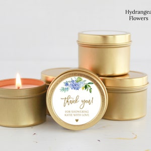 May include: A group of round gold metal candle tins. One tin is lit with a visible flame. A circular white label on the front features a watercolor hydrangea illustration, the text "thank you!" and "FOR SHOWERING KATIE WITH LOVE" with a small heart.